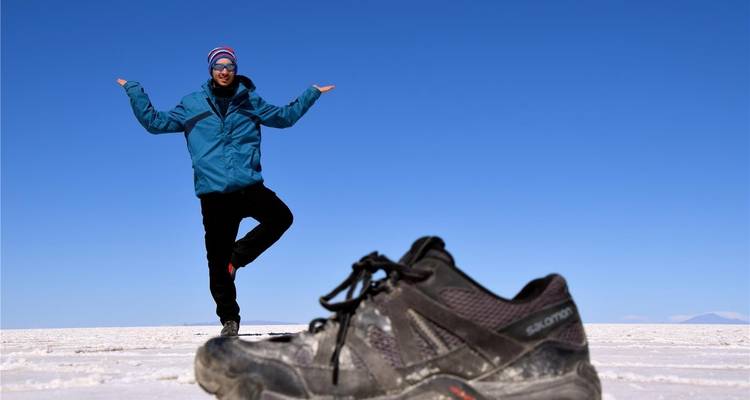 Perspektivtrick-Foto eines Mannes, der neben einem übergroßen Schuh auf den weitläufigen weißen Salzseen von Uyuni unter blauem Himmel balanciert.