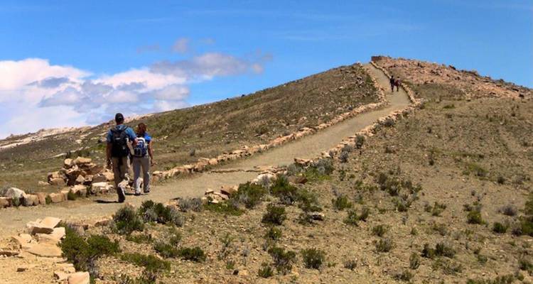 Los excursionistas caminan por un sendero bordeado de piedra atravesando una cresta árida bajo un cielo azul brillante.