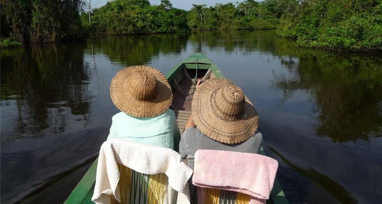 Two passengers wearing straw hats sit at the front of a green boat cruising a calm river in the jungle.