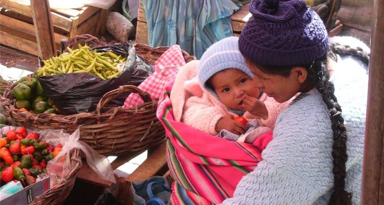 Indigene Frau wiegt ihr Baby in den Armen, während sie farbenfrohe Produkte auf einem belebten Freiluftmarkt verkauft.