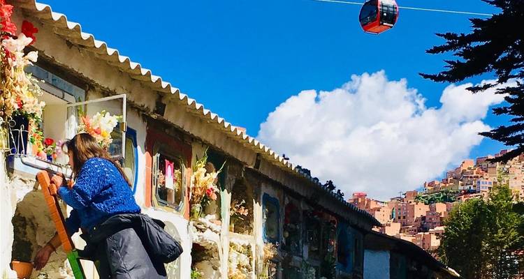 Woman decorating a colourful cemetery wall as a red cable car travels overhead toward hillside homes.