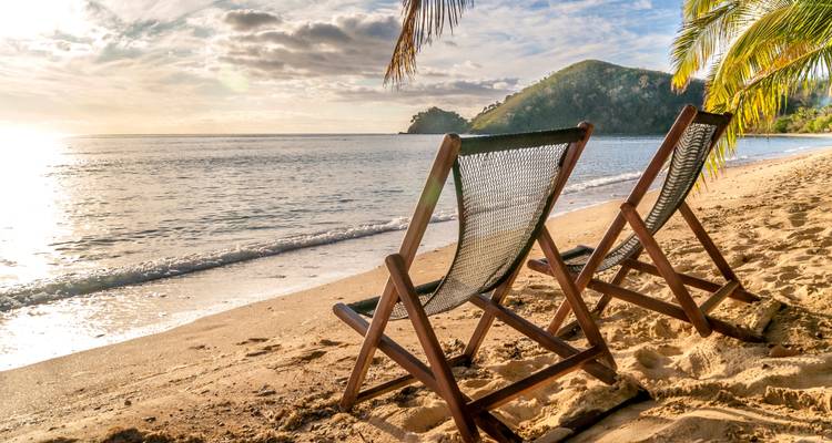 Two empty wooden deck chairs face a calm sea on a sandy beach in warm late-afternoon light.