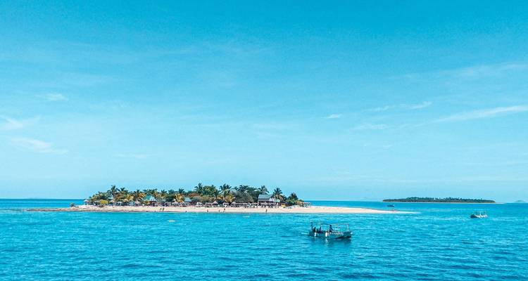A tiny palm-lined island sits in vivid turquoise waters with a couple of small boats floating nearby under a clear sky.