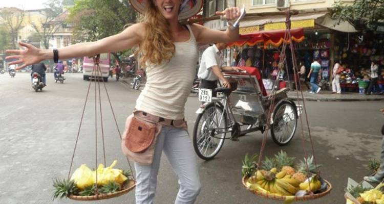 Femme souriante portant un chapeau conique qui transporte de manière ludique des paniers de fruits tropicaux sur une rue animée de Hanoi.