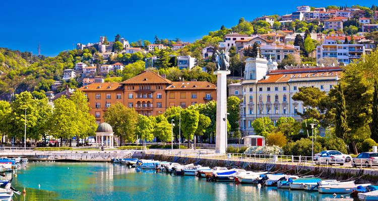 Malerische Marina mit an türkisblauem Wasser vertauten Booten und farbenfrohen Gebäuden, die sich an einem grünen Berghang in Rijeka erheben.