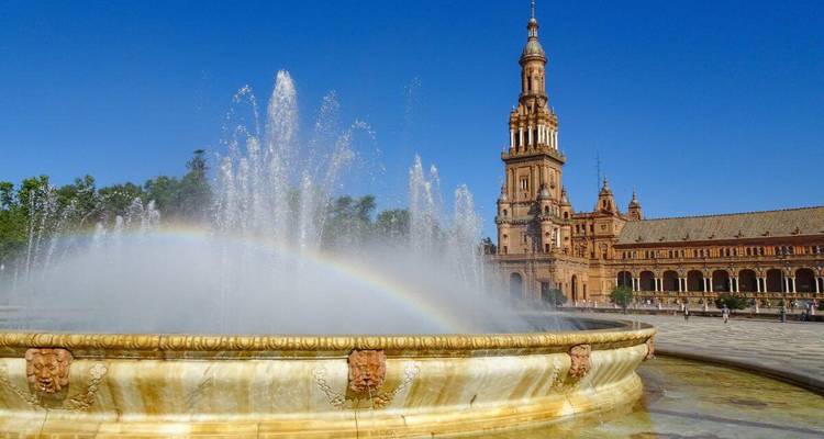 Weitaufnahme des Brunnens der Plaza de España mit einem Regenbogenbogen und dem verziert gestalteten Ziegelturm vor einem klaren blauen Himmel