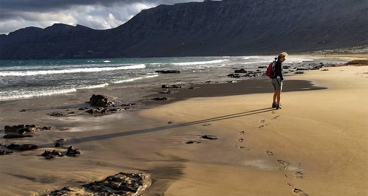 Un excursionista solitario con mochila camina por una playa dorada vacía, dejando huellas hacia el oleaje ondulante y acantilados oscuros.