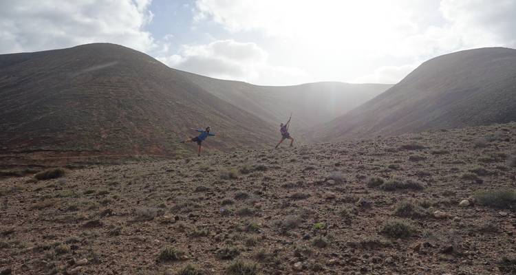 Dos excursionistas saltando alegremente en una ladera volcánica árida entre colinas redondeadas con una fuerte contraluz.
