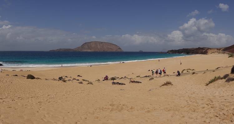Playa de arena dorada y ancha con océano color turquesa e isla frente a la costa, con bañistas dispersos disfrutando del clima despejado.