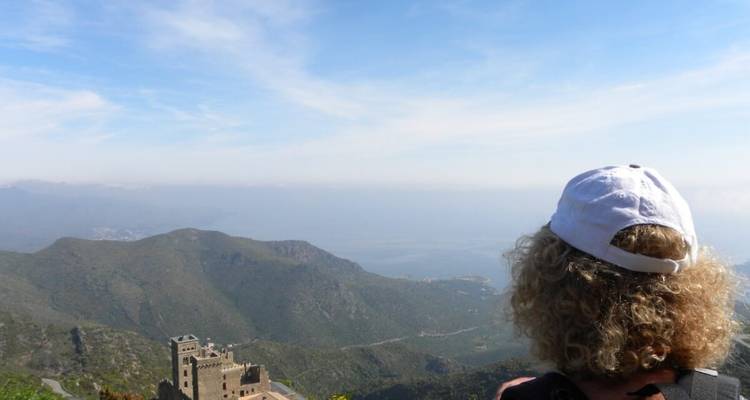 Un voyageur avec une casquette blanche regarde au loin par-dessus des montagnes côtières escarpées et un château en contrebas.