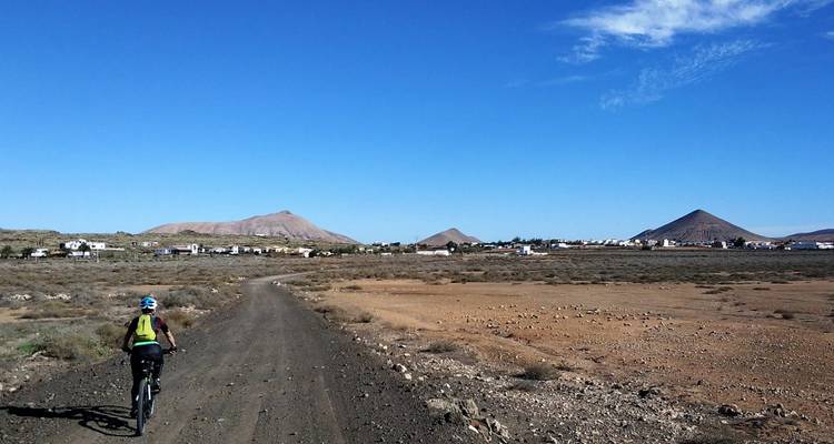 Cyclist rides a rough dirt road across a barren volcanic plain toward cone-shaped hills.