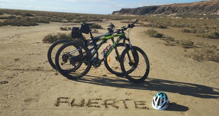 Two mountain bikes rest on sandy ground where the word 'FUERTE' is written beside a helmet.