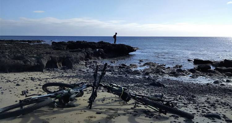 Silhouetted figure stands on volcanic rocks beside a tranquil sea while bikes lie on the sand.