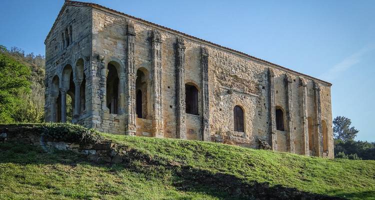Un bâtiment historique en pierre avec des fenêtres cintrées situé sur une colline verdoyante sous un ciel dégagé.