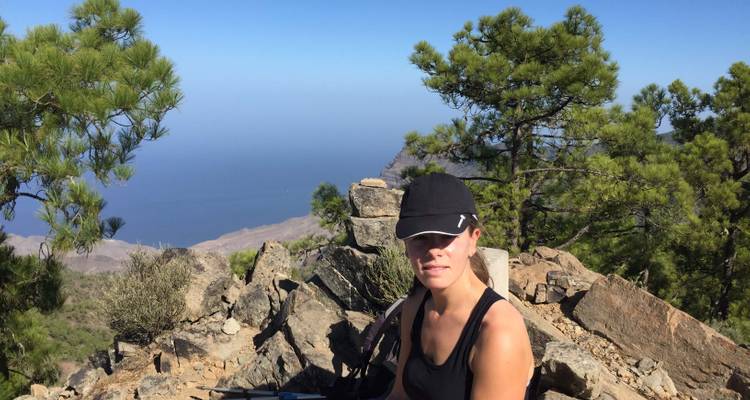 Personne assise sur des rochers avec vue sur les arbres et la mer au loin.