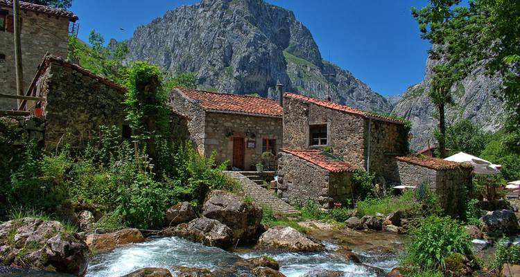 Ein rustikales Steinhaus mit rotem Dach an einem Bergbach, umgeben von Grün.