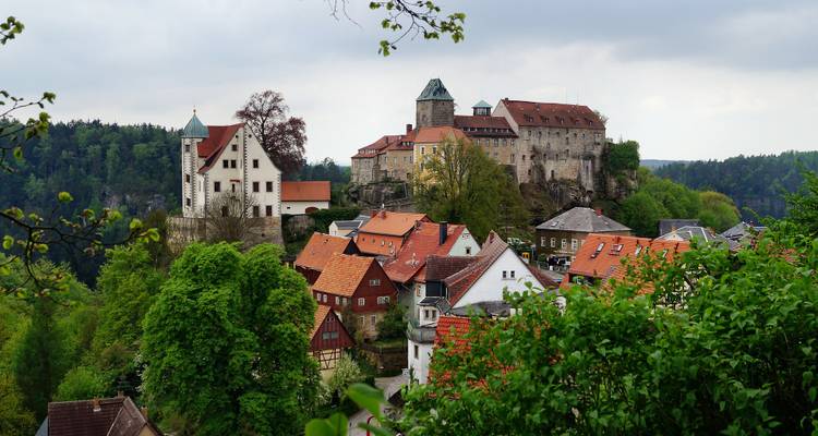 Château médiéval au sommet d'une colline avec des maisons aux toits rouges et un paysage verdoyant.