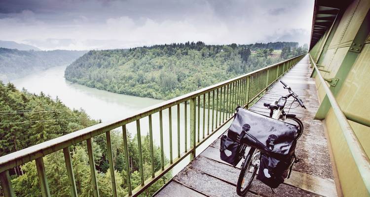 Vélo de randonnée chargé sur un pont étroit surplombant une vallée fluviale brumeuse et une forêt