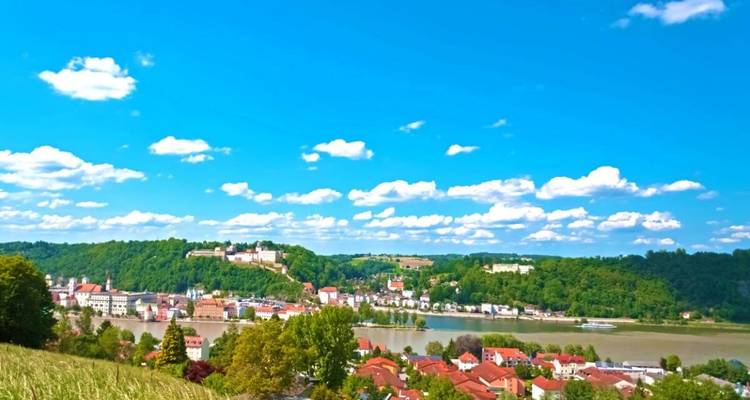 Landschaftlicher Blick auf einen Fluss, der von einer kleinen europäischen Stadt und üppigen Hügeln gesäumt wird.