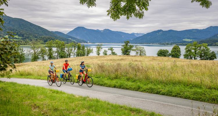 Drie fietsers trappen langs een meer-zijweide onder een bewolkte hemel met alpenbergen aan de overkant van het water.
