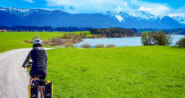 Fietser rijdt over een grindpad naast een rustig meer met besneeuwde Alpen die daarachter hoog oprijzen.