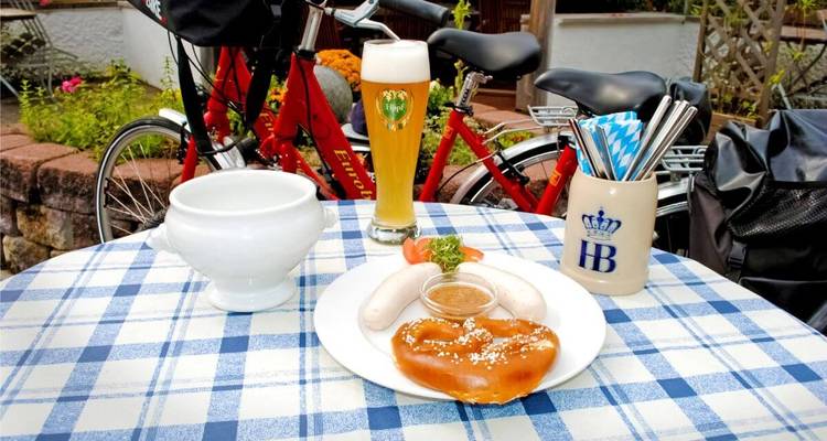 Mesa con un desayuno alemán que incluye pretzel, salchichas y bebida, con una bicicleta en el fondo.
