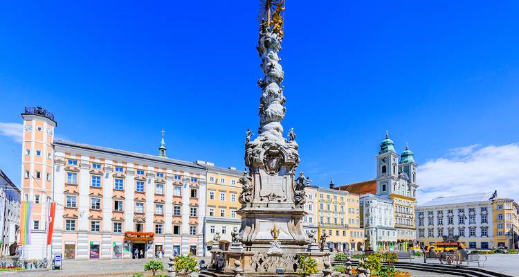 Lebendiger Stadtplatz mit historischen Gebäuden und verzierter Säule.