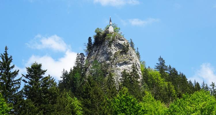 Een scherpe rotsige bergtop gekroond met een kleine kapel rijst op boven dicht groen bos tegen een heldere blauwe hemel.