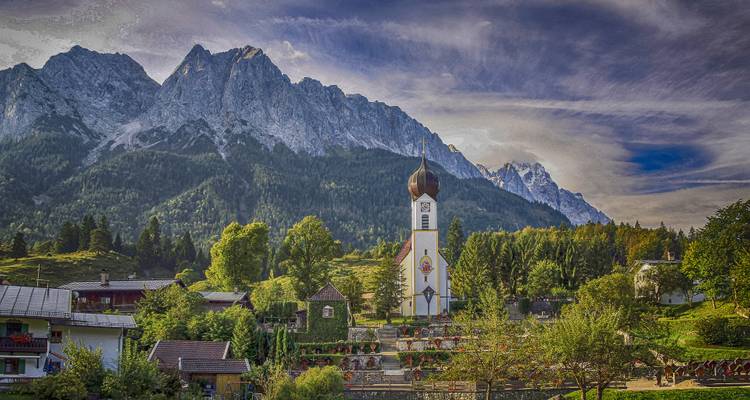 Dorp met een kerk en alpine bergachtergrond onder een dramatische lucht.
