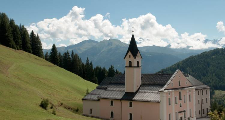 Kirche auf einem grasbewachsenen Hügel mit Bergkulisse unter blauem Himmel.