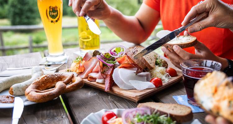 Traditionelles bayerisches Picknick mit Fleisch, Brezel, Käse und Bier auf rustikalem Holztisch.