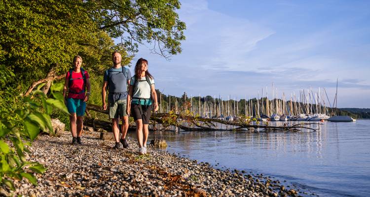 Wandelaars lopen langs de kiezelstrand van het meer bij een jachthaven vol zeilboten in zacht avondlicht.