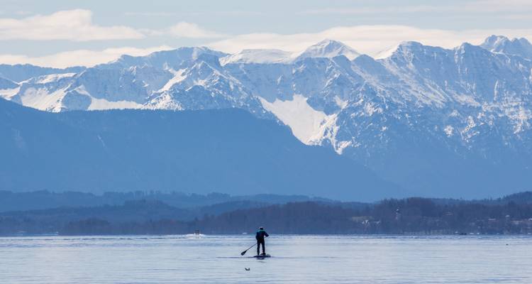 Person beim Paddleboarding auf einem See mit schneebedeckten Bergen im Hintergrund.