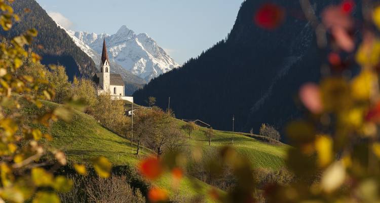 Eine kleine Kirche auf einem Hügel mit schneebedeckten Bergen im Hintergrund, umgeben von Bäumen.