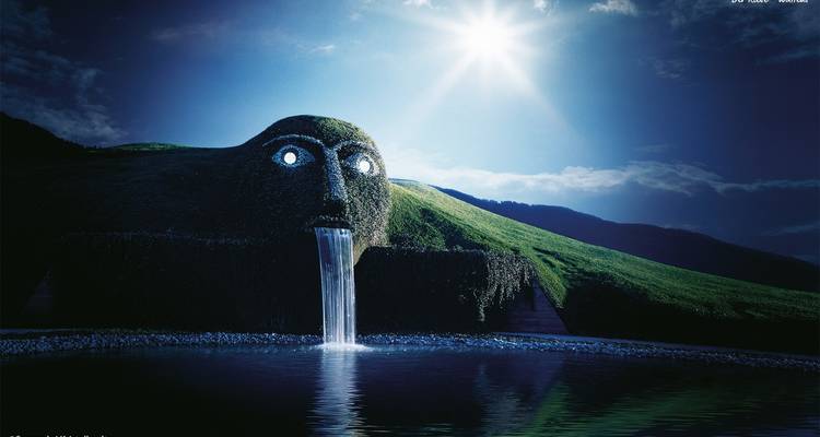Artistic installation of a giant head sculpture spouting water into a pool, under a bright sky.