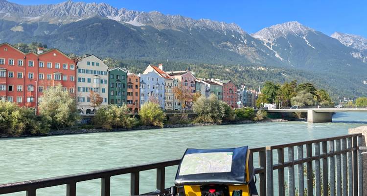 Un vélo rouge EuroBike est appuyé contre une rambarde surplombant les maisons colorées en rangée d'Innsbruck au bord de la rivière Inn aux flots rapides avec les sommets alpins en arrière-plan.