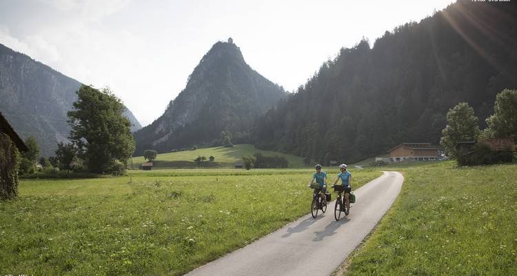 Deux cyclistes roulant sur un sentier à travers de luxuriants champs verts avec des montagnes en arrière-plan.