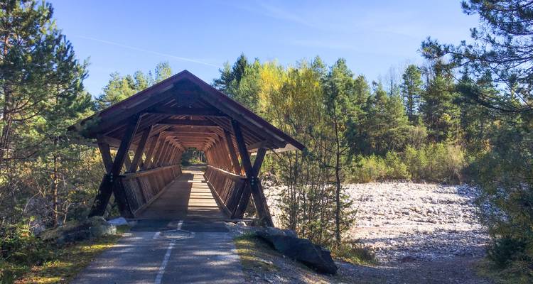 Eine überdachte Holzbrücke über einem steinigen Flussbett mit umgebenden Bäumen.