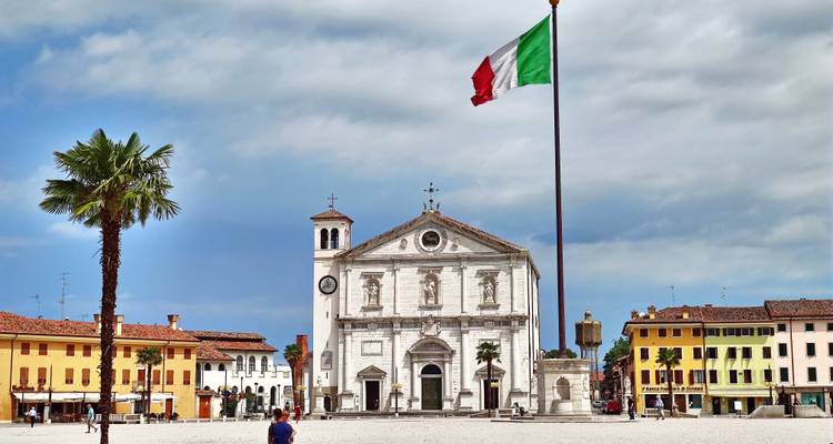 Ein großartiger italienischer Platz mit zentraler Kirche und hohem Fahnenmast, umrahmt von bunten Gebäuden und Palmen.