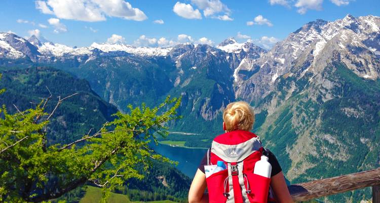 Wanderer mit rotem Rucksack blickt auf eine Bergaussicht mit einem See darunter.