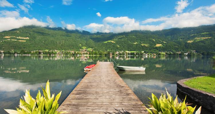 Houten steiger die zich uitstrekt in een spiegelglad alpenmeertje met kleine bootjes en beboste bergen.