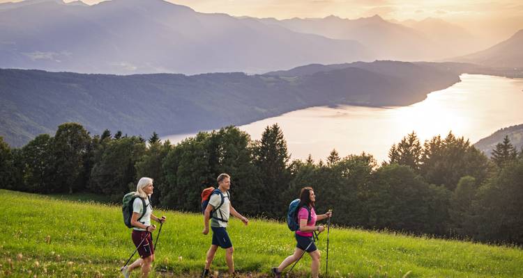 Drie wandelaars met rugzakken lopen door een bloemrijke weide met uitzicht op een zonbeschenen meervallei.