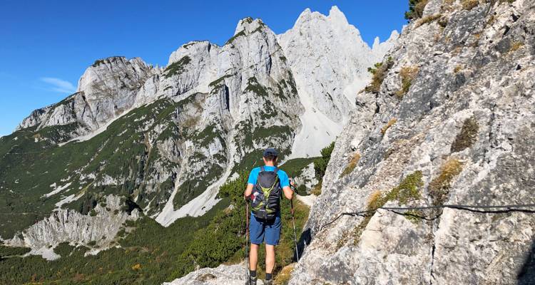 Solo hiker on a rocky alpine trail below towering white limestone cliffs.
