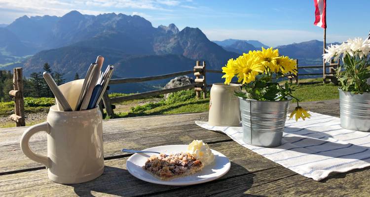 Rustic wooden table on mountain hut terrace with cake, flowers and distant peaks.