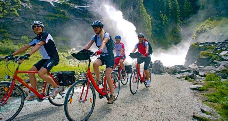 Group of cyclists riding past a waterfall surrounded by greenery.