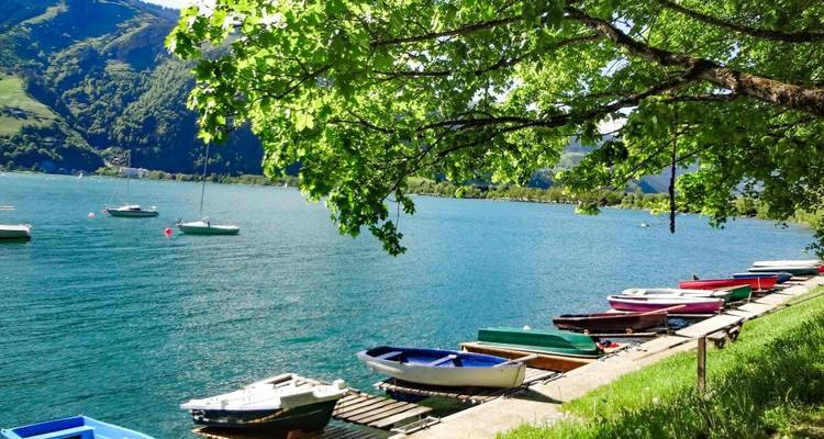 Boats docked on a lake with a mountainous backdrop and tree-lined path.