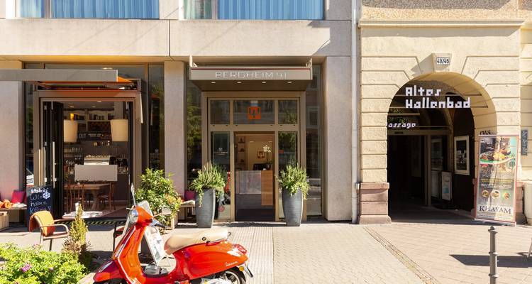 Straßenansicht eines modernen Cafés, Boutique-Hotel-Eingang und der historischen Altes Hallenbad Arkade in Heidelberg mit einem roten Roller, der draußen geparkt ist.