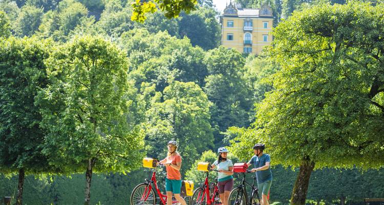 Cyclists with bikes near trees and a large yellow building in the background.