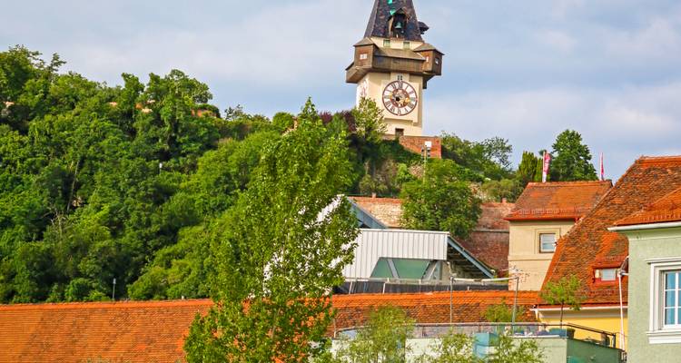 Graz' berühmter Uhrturm erhebt sich über rotgedeckte Dächer und grüne Hügelhänge.