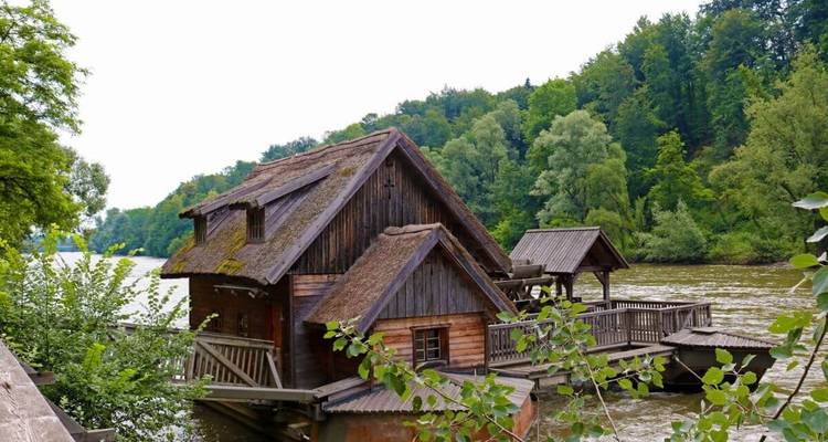 A charming wooden water mill on a riverbank surrounded by trees.